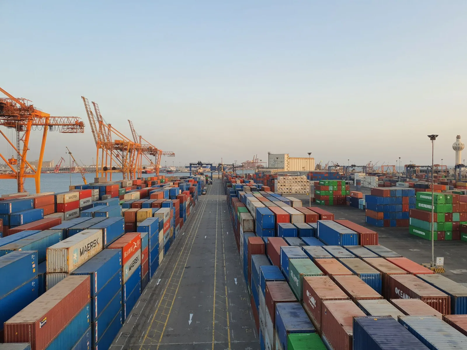 Rows of stacked shipping containers at a port with cranes in the distance